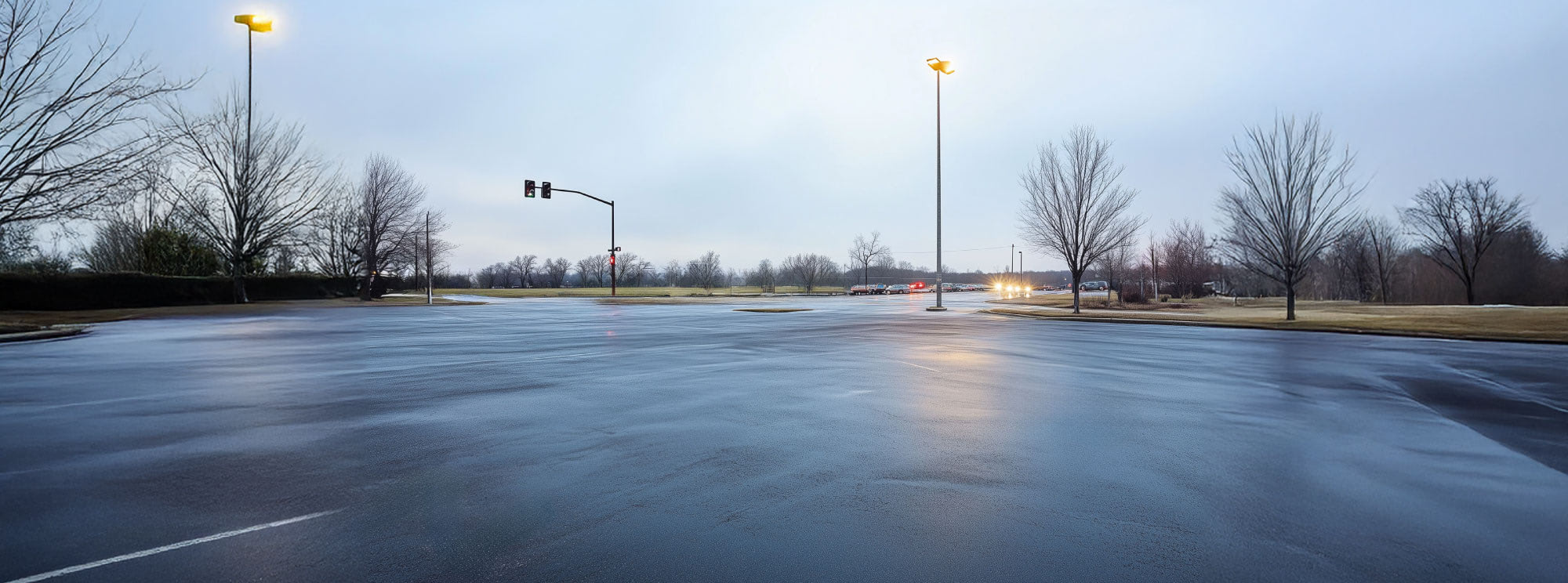 An empty parking lot, glistens from a light rain. Lights glow dimly in the early evening light.